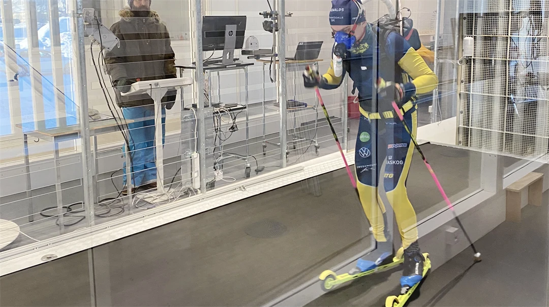 Man i blågul dräkt på rullskidor i vintunneln. En annan man står utanför vindtunneln.