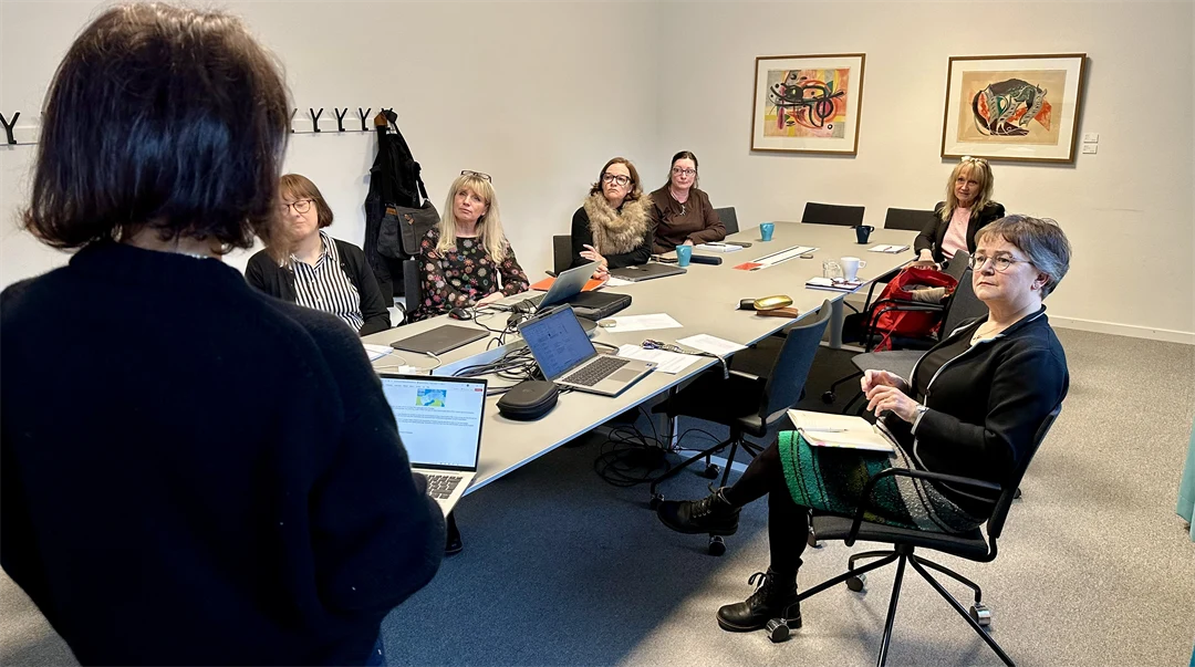 Sex kvinnor sitter runt ett konferensbord och tittar mot en annan kvinna som står upp med ryggen mot kameran.