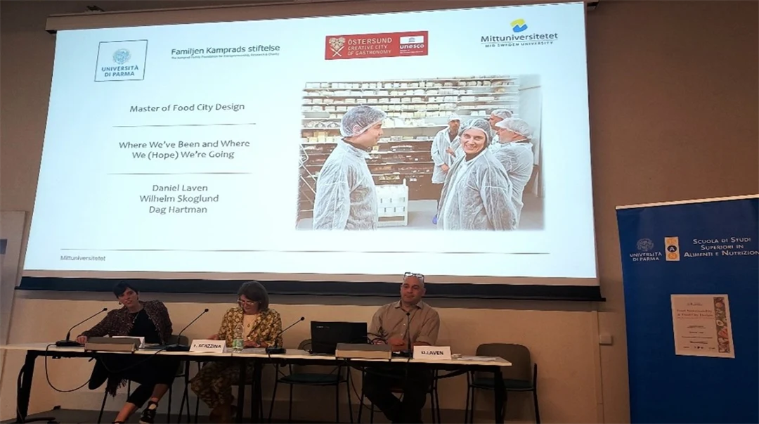 Three people sitting at a table with computers in front of them and a large screen behind them, presenting at a conference