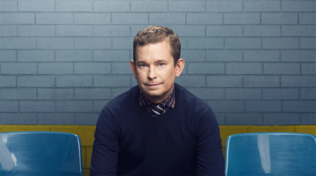 En man sitter på en stol i en stolsrad framför en blå och gul vägg.