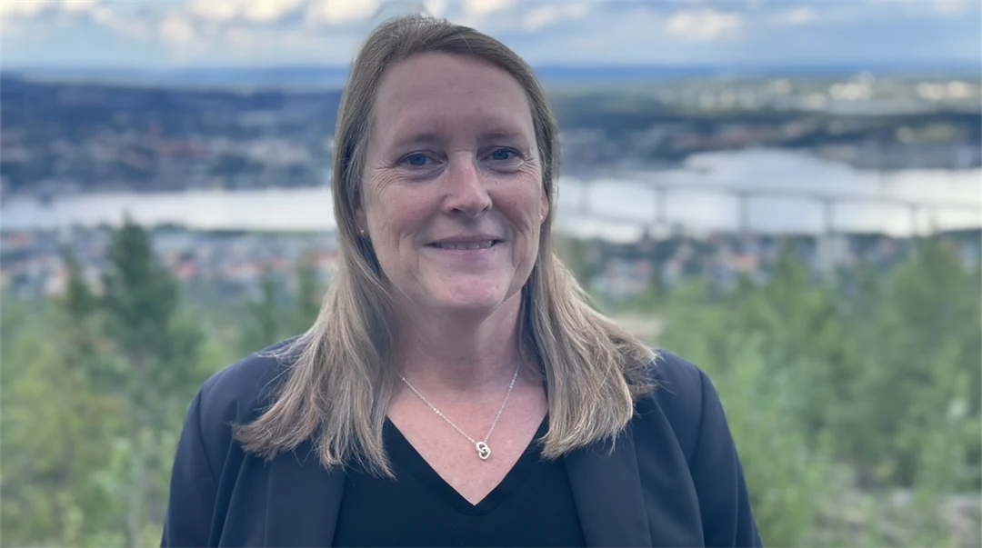 En kvinna med blont axellångt hår, svart tröja och mörkblå kavaj med en naturskön vy med skog, hav och en bro i bakgrunden.