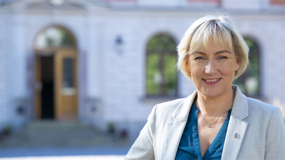 En kvinna i grå kavaj och blå blus står framför en byggnad.