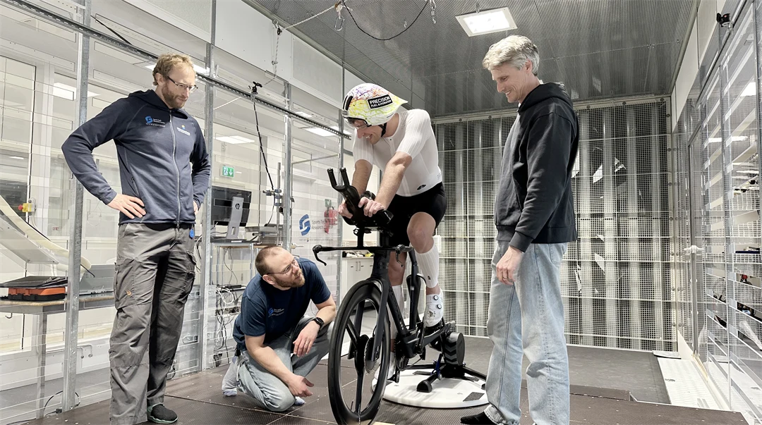 Tre män står och tittar på en fjärde man som sitter på en cykel inomhus.