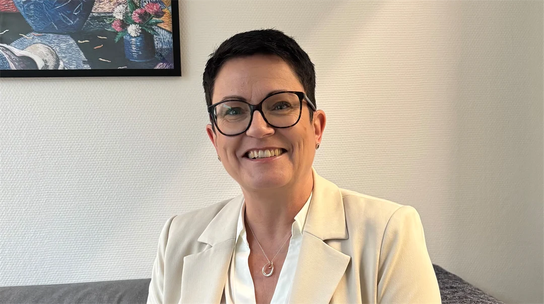 En kvinna med mörkt kort hår, klädd i cremevit kavaj och vit blus sitter i en soffa med en tavla på väggen bakom sig. 