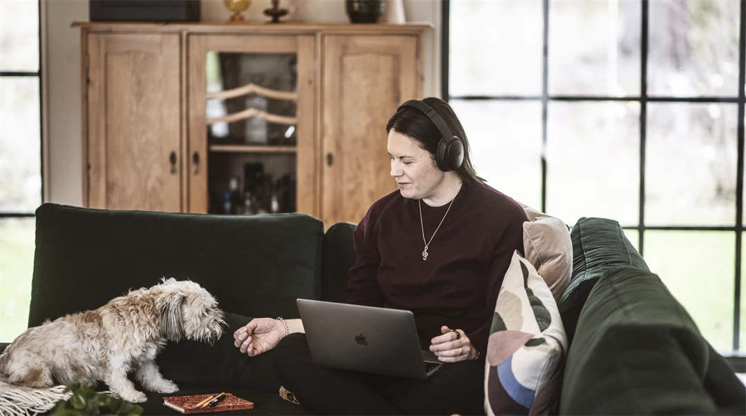 En kvinna sitter i en soffa, med hörlurar på huvudet och en dator i knät. 