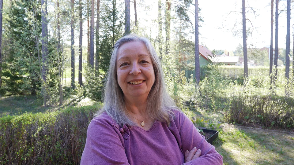 En gråhårig kvinna i lila tröja står i en trädgård och ler mot kameran.