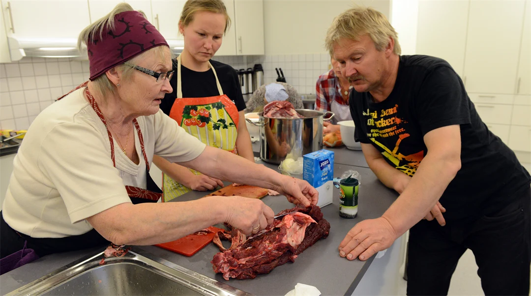 En äldre kvinna visar en ung kvinna och en man hur man tillagar renkött.