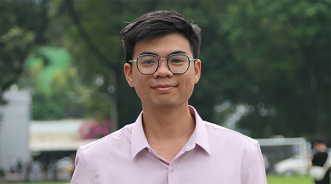 A young man in a pink shirt and glasses is smiling and looking into the camera.