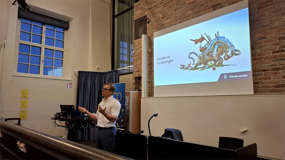 en man i vit skjorta står i en föreläsningssal framför en stor bildskärm