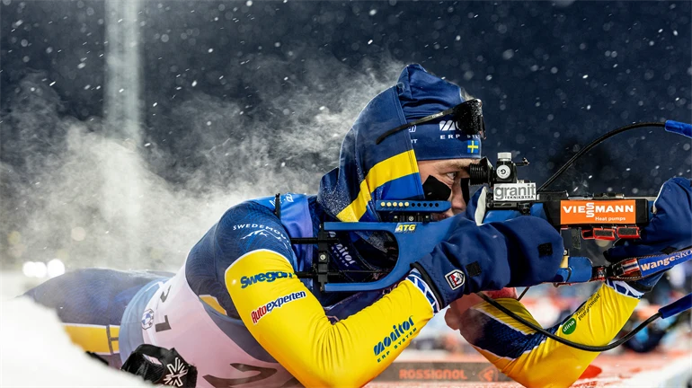 Biathlonist lying down aiming his rifle, steam coming from his back.