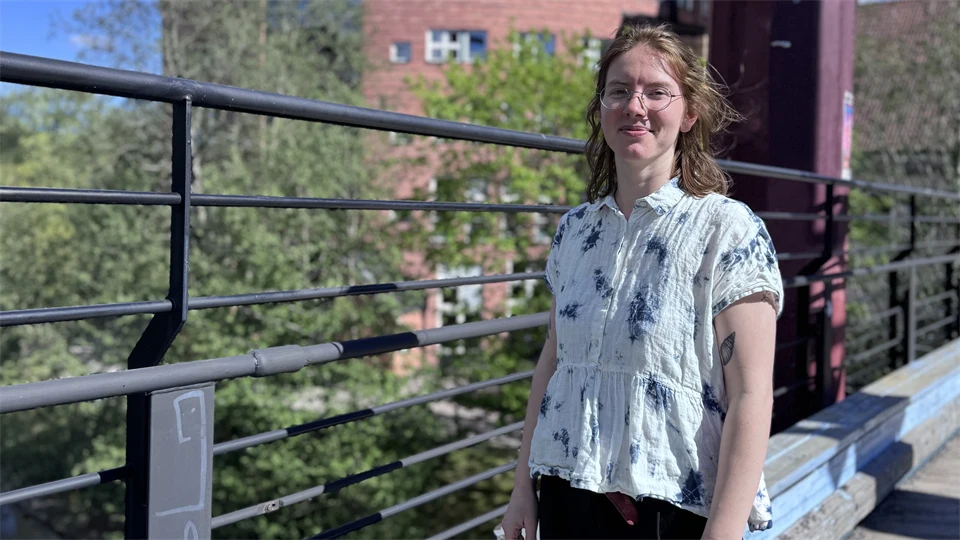 A student is smiling. In the background the campus is visable. 