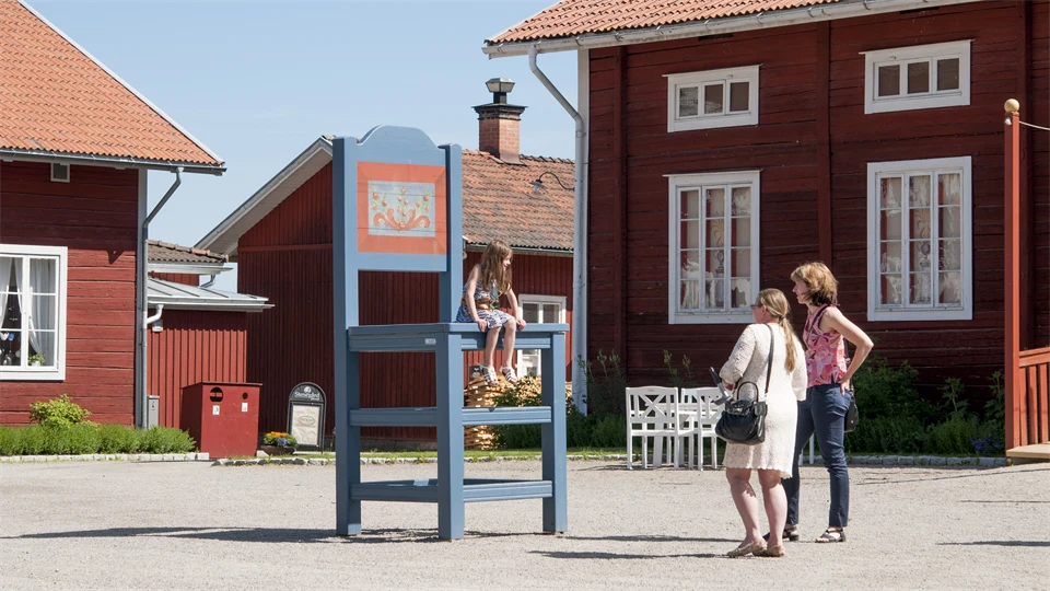 A girl sits on a large chair outside three red houses and two women stand below and look at the girl.