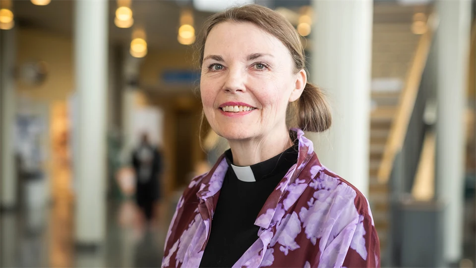 Kvinna med svart skjorta med prästkrage ler mot kameran. 
