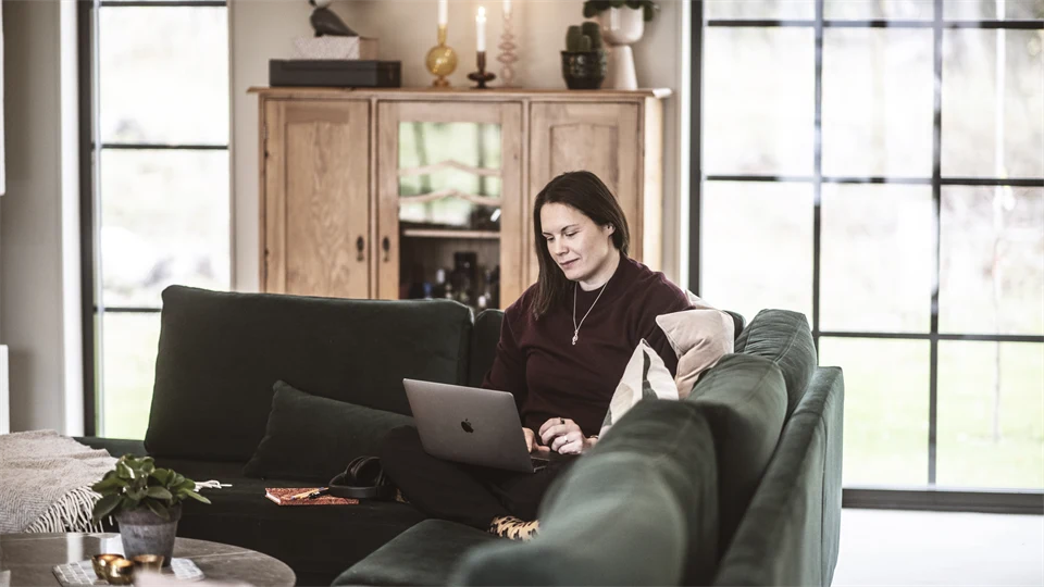 En kvinna sitter i soffan och jobbar med sin dator i knät. 