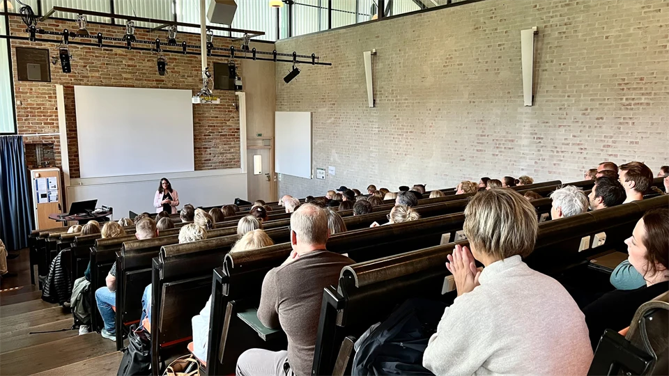 En föreläsningssal med ett femtiotal deltagare som sitter på sina stolar och lyssnar på en kvinna som pratar längst fram i salen.