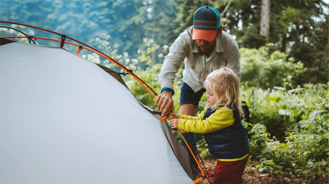 Ung man och hans dotter tre-årsåldern sätter upp ett tält tillsammans i skogen. De campar och är klädda i friluftskläder.