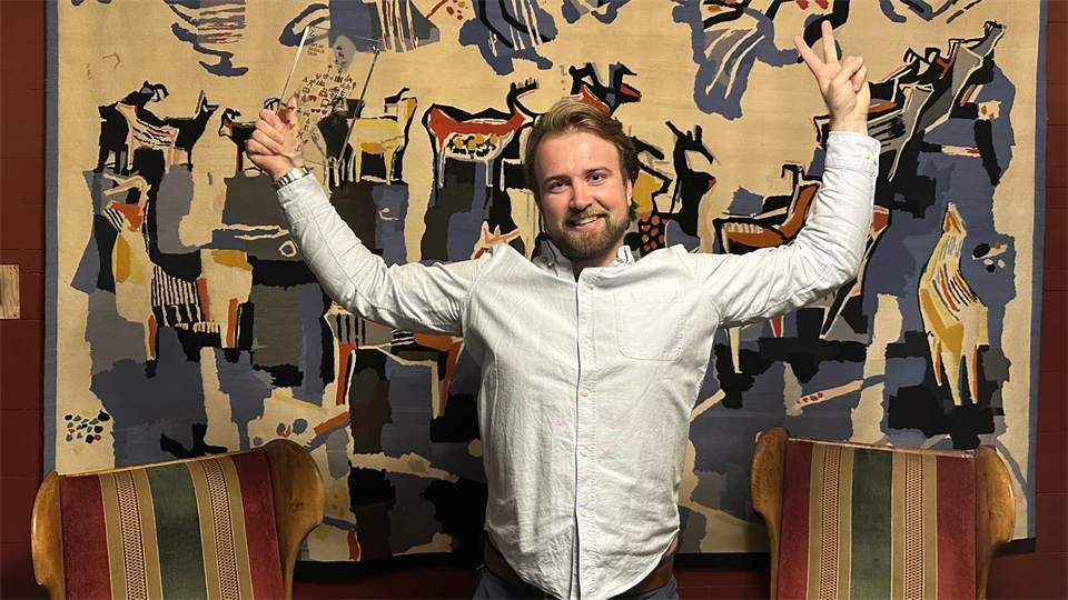 Man i vit skjorta står framför en bonad med händerna uppsträckta i luften. I den ena handen håller han ett pris i glas. 