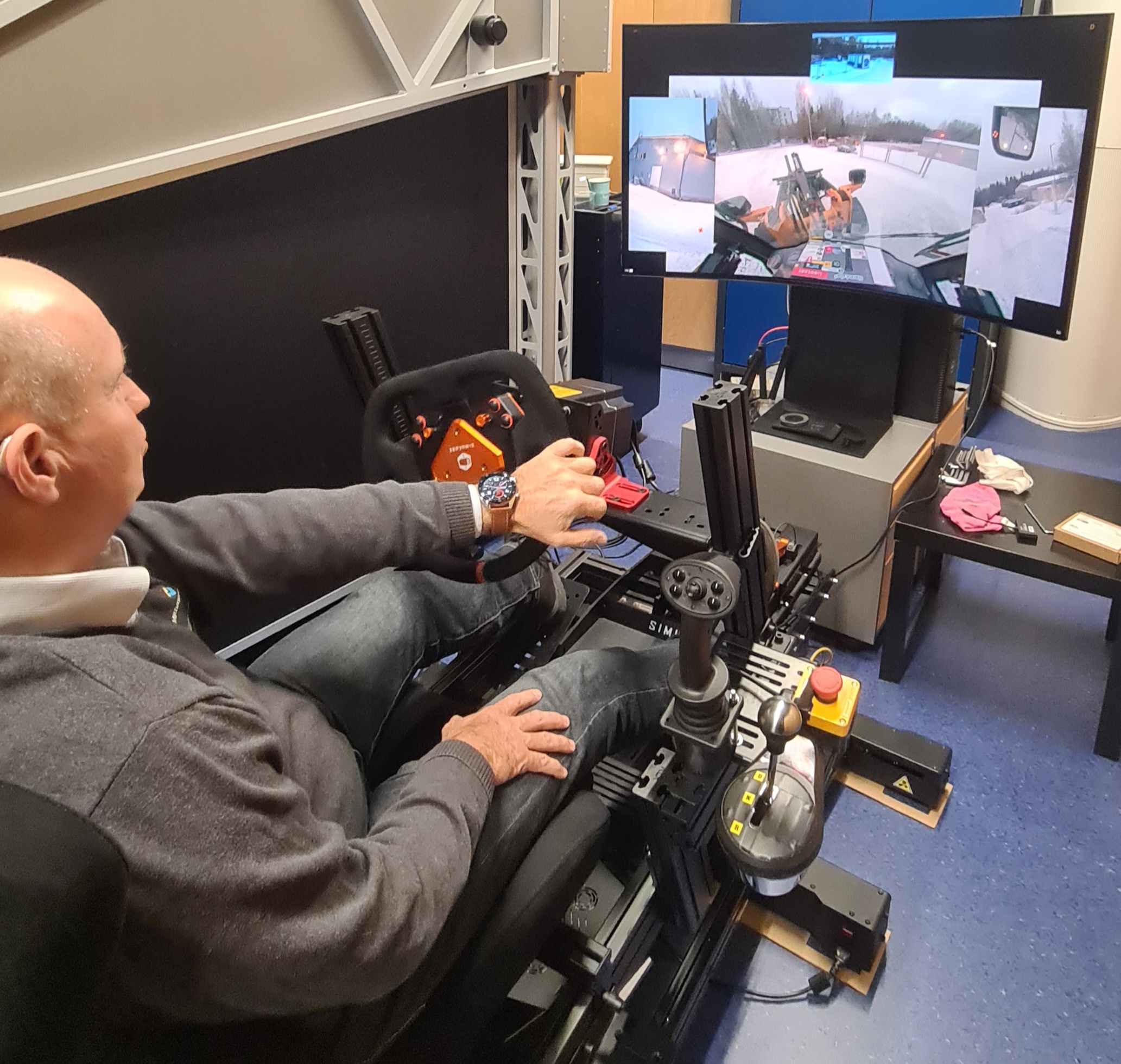 En man sitter i en förarstol med ratt och tittar på en skärm som presenterar en förarvy