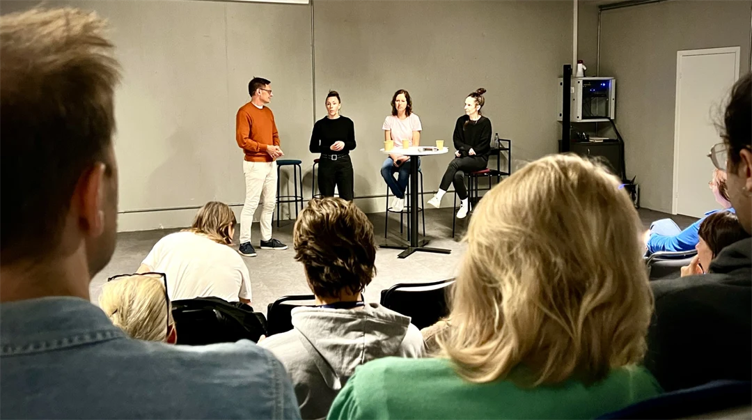 Fyra personer står vid ett runt bord och pratar fram mot en publik.