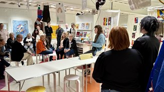 En kvinna sitter på ett bord framför en publik på runt ett tjugotal deltagare.