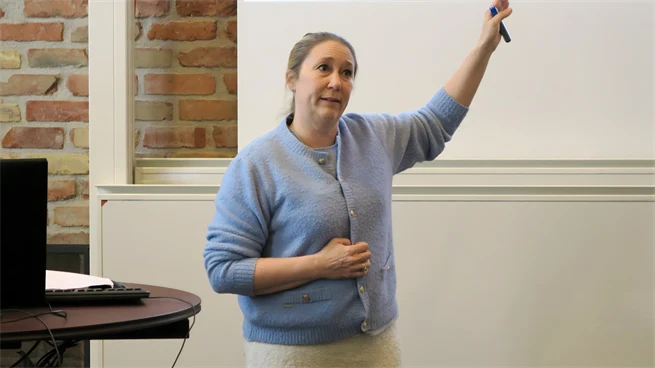 Kvinna står längst fram i en aula och pratar med åhörarna. Hon har ljust hår och bär en ljusblå kofta. 