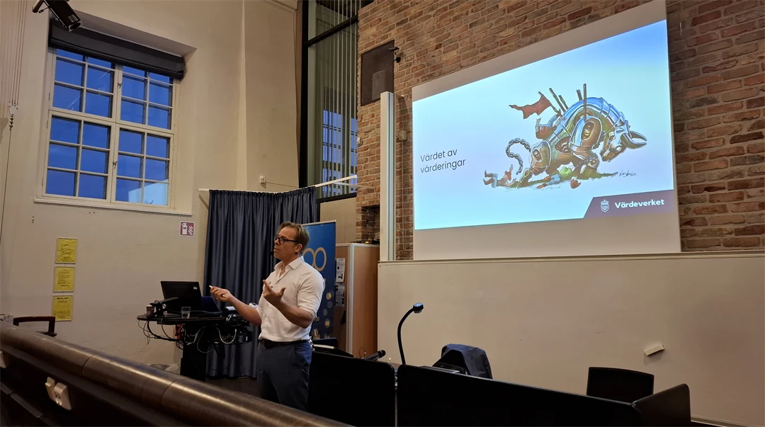 en man i vit skjorta står i en föreläsningssal framför en stor bildskärm