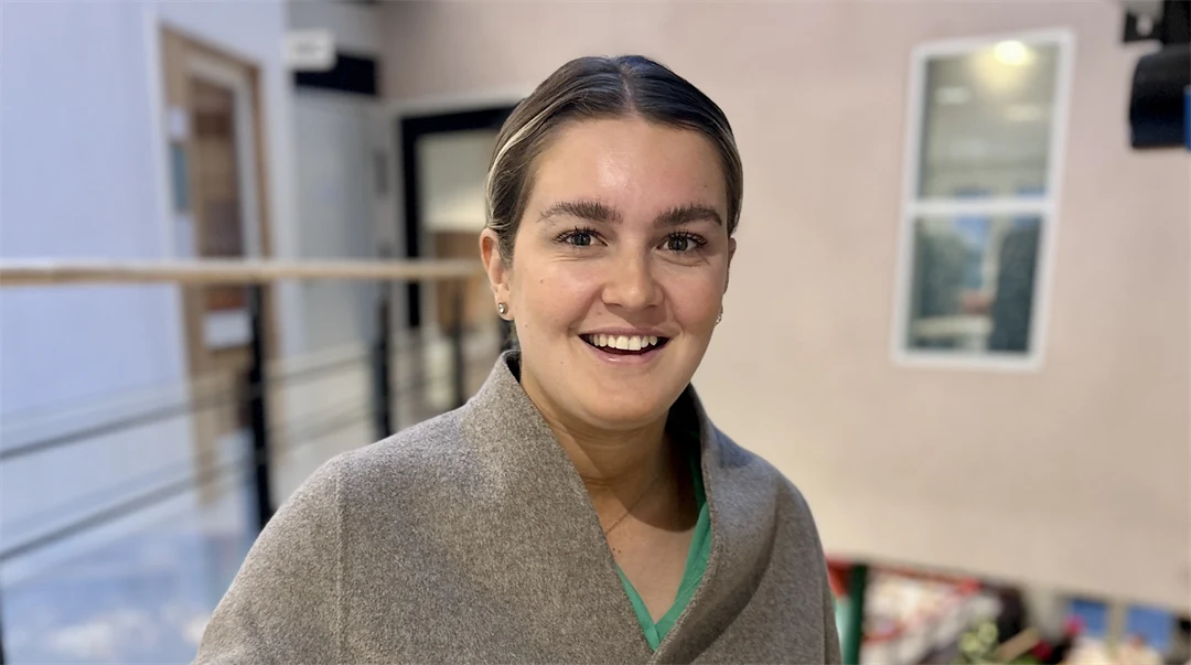 En mörkblond kvinna klädd i beige kofta står i trappa med beige vägg bakom sig.