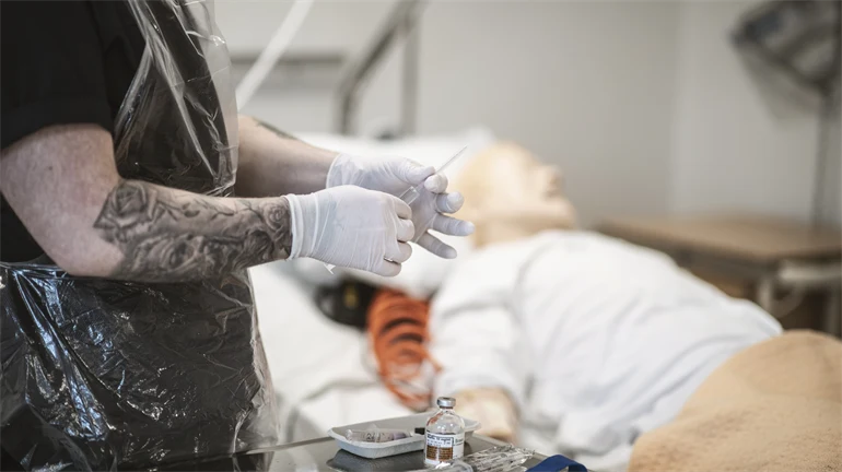 En man med skägg är i en sjukhusmiljö och övar med en spruta på en patientdocka.