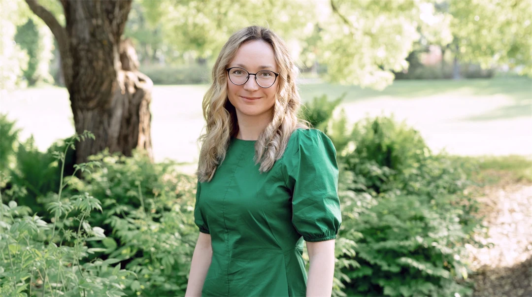 Ung kvinna med mellanlångt ljust hår och glasögon. Hon bär en grön klänning och står i en grönskande trädgård.