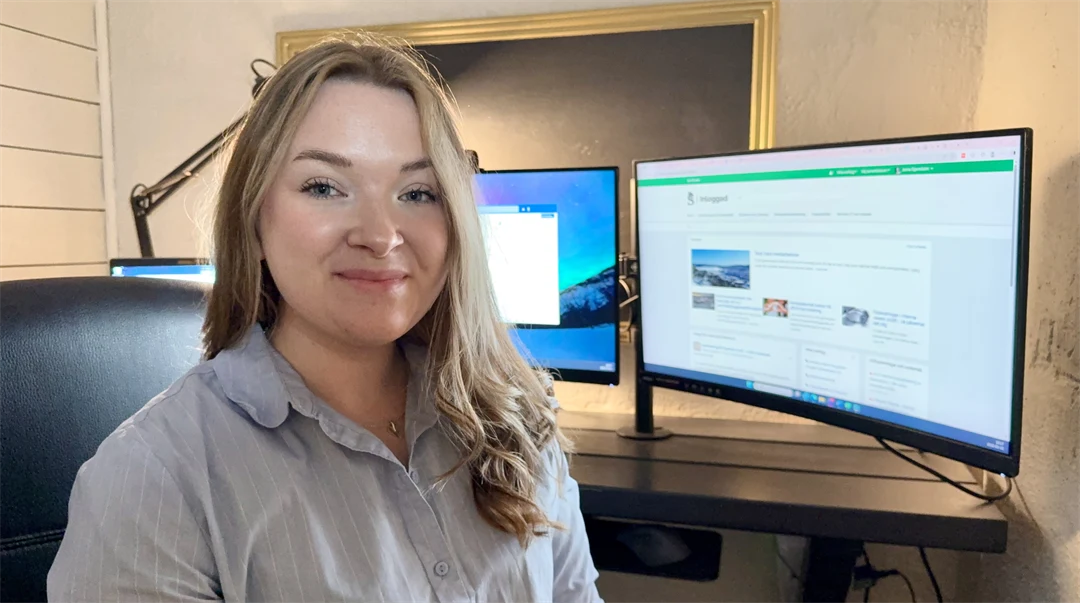 En kvinna tittar in i kameran och ler. I bakgrunden står två dataskärmar.