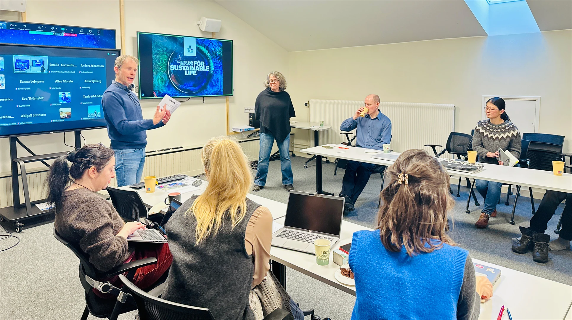 En man står längst fram i ett rum framför två skärmar och tittar ut över salen med åhörare medan han håller i ett papper och pratar.