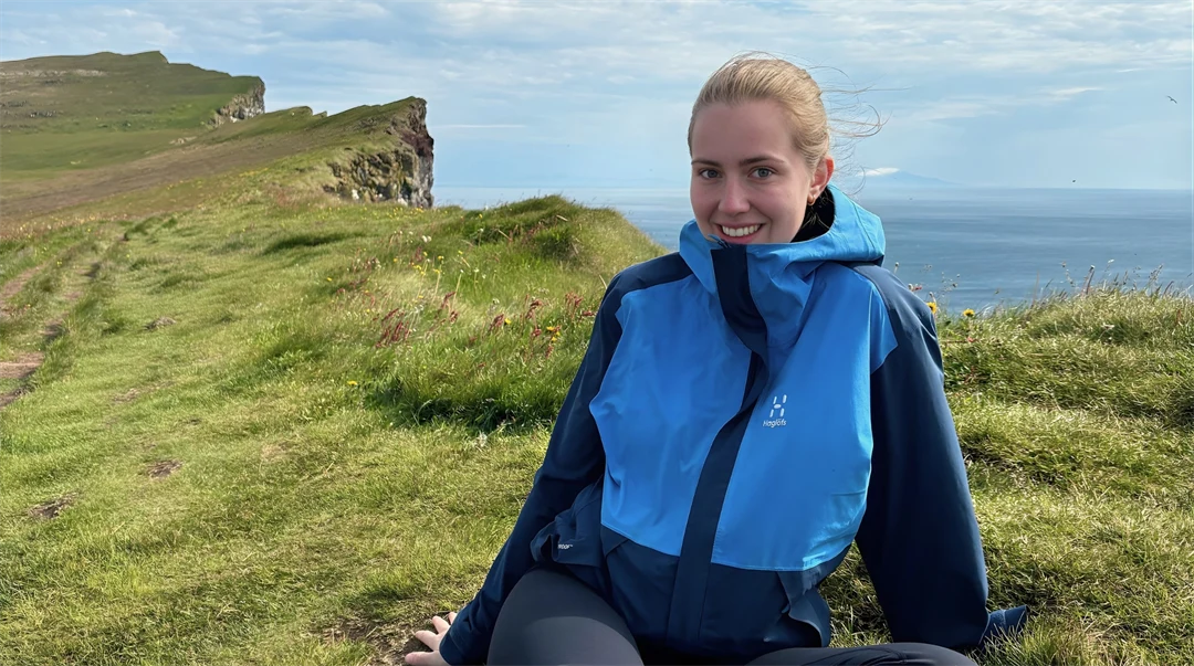 Ung kvinna i blå vindjacka, sittandes på en bergskam med havet nedanför. Hon är glad.