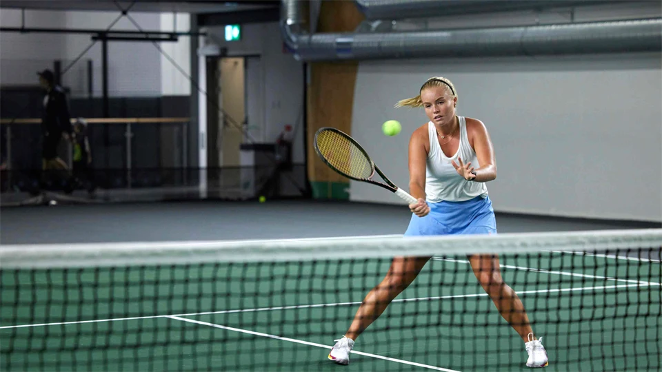 Kvinna som spelar tennis inomhus, hon står vid nät och ska till att slå en volley.