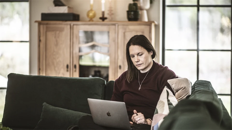 En kvinna sitter i soffan och jobbar med sin dator i knät. 