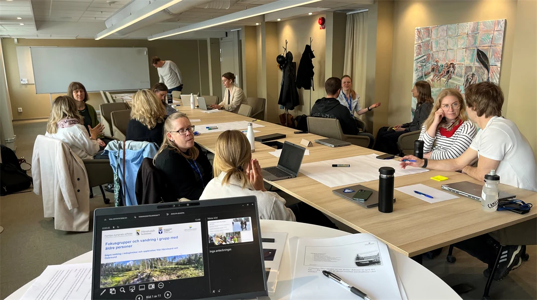 Ett tiotal personer sitter runt ett bord och pratar med varandra och tittar på en skärm med en presentation