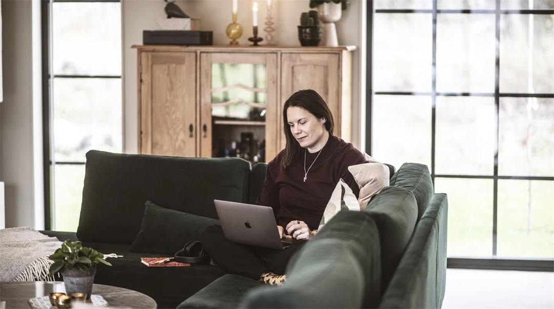 En kvinna sitter i soffan och jobbar med sin dator i knät. 