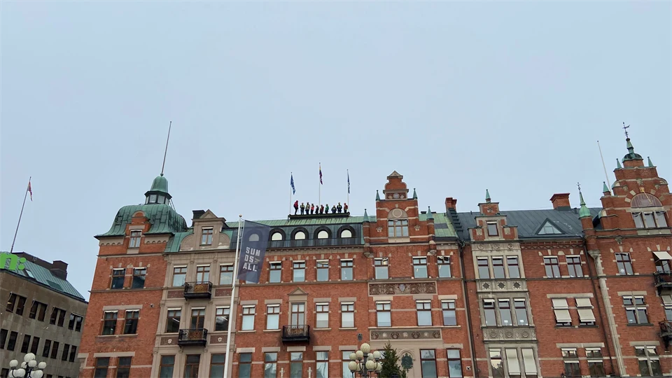 En grupp människor står på ett hustak. Huset är en pampig tegelbyggnad med valv och torn.