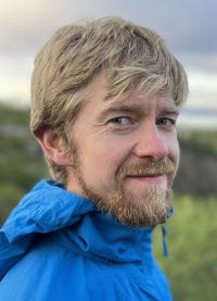 En man i ljust hår, skägg och blå jacka i funktionsmaterial står utomhus med ett grönt naturlandskap i bakgrunden.