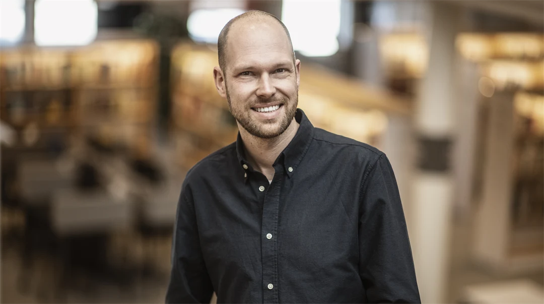 En man i svart skjorta med vita knappar ler mot kameran