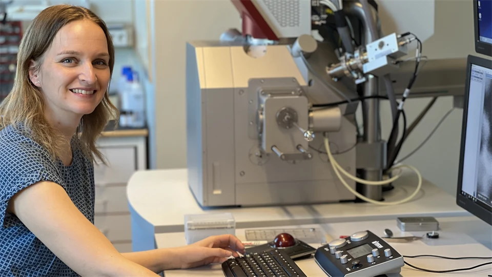 A woman sits at a desk with advanced equipment beside her. She looks into the camera and smiles.