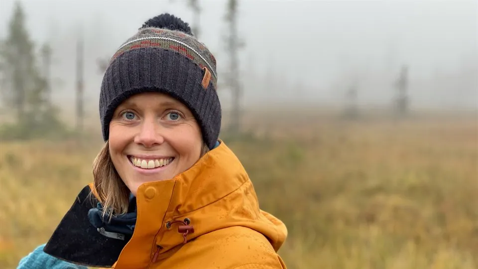 Sara Häggström står på en öppen plats i skogen med några träd i bakgrunden. Det är dimma bakom henne. Sara har på sig en orange jacka och en stickad mössa. Hon ser in i kameran och ler brett. 