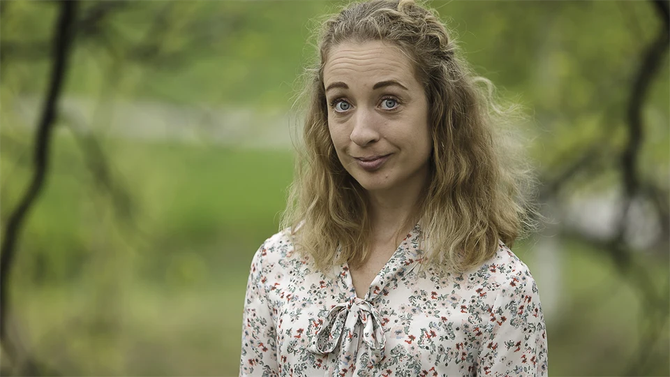 Woman with curly shoulder-length hair and floral blouse standing outdoors with green trees around her.