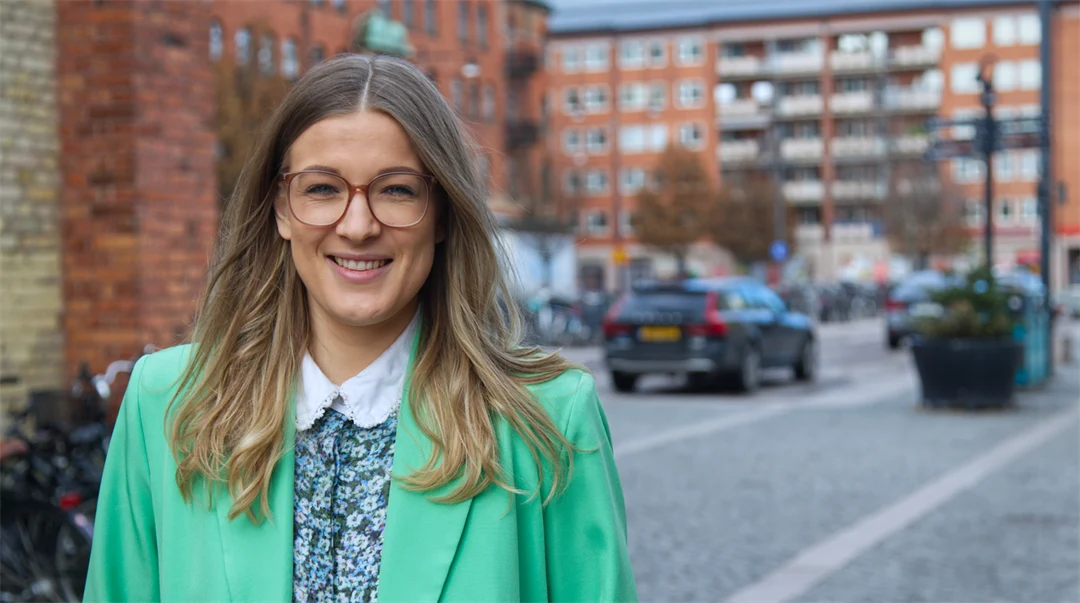 En kvinna i en grön kavaj står framför några byggnader.