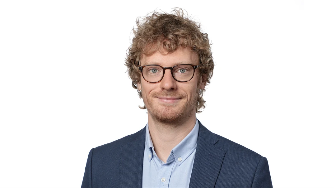A man with curly hair, glasses, wearing a light blue shirt and dark blue jacket.