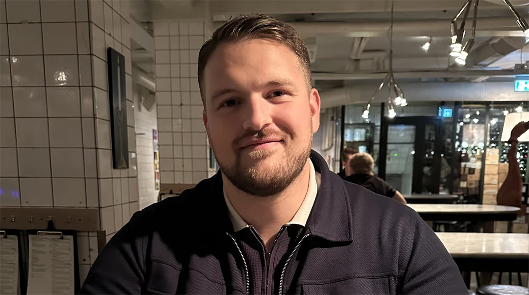 Man med mörkt kort hår, skägg och mörkblå kofta tittar in i kameran.