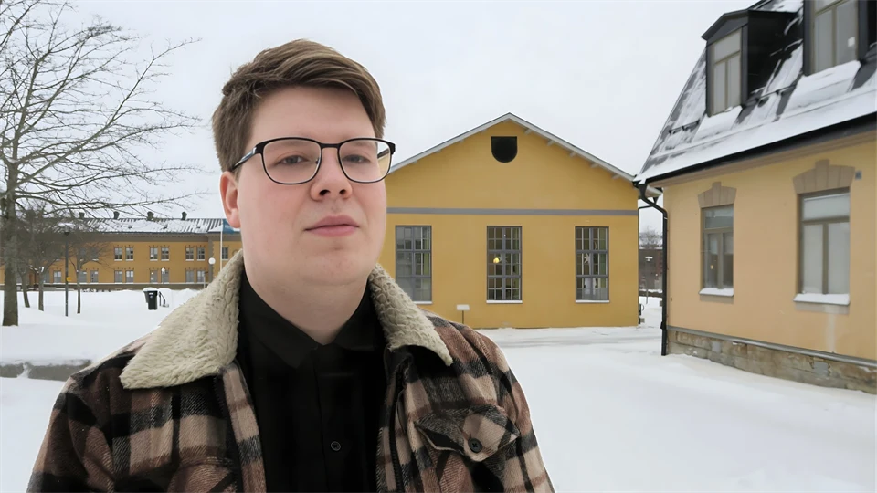 Manlig student står på campusgården i Östersund framför några av byggnaderna. Gården är snötäckt och studenten har vinterjacka på sig.