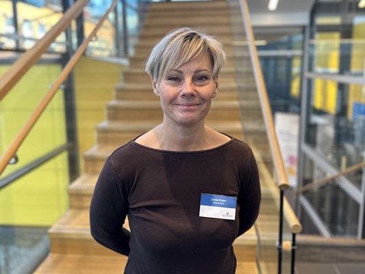 En kvinna med kort blont hår, brun tröja och namnskylt står framför en trappa med glaspartier och gulgröna detaljer runt om kring.