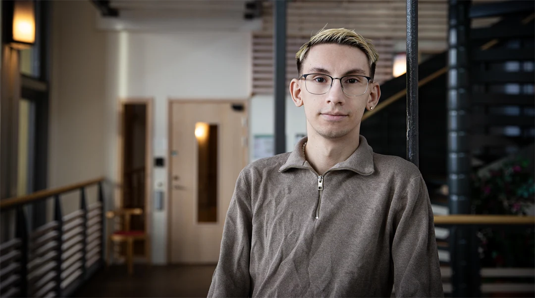 En man med glasögon och beige tröja står i en korridor.