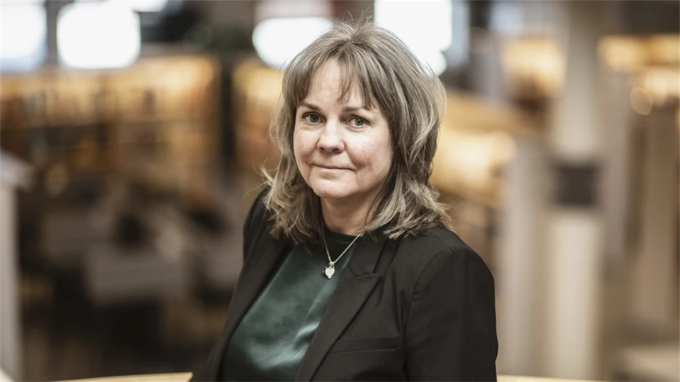 En kvinna i ljusbrunt hår, grön blus och svart kavaj står i biblioteksmiljö.