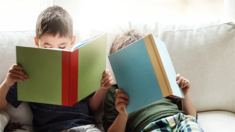 Two boys are sitting on a sofa reading books.